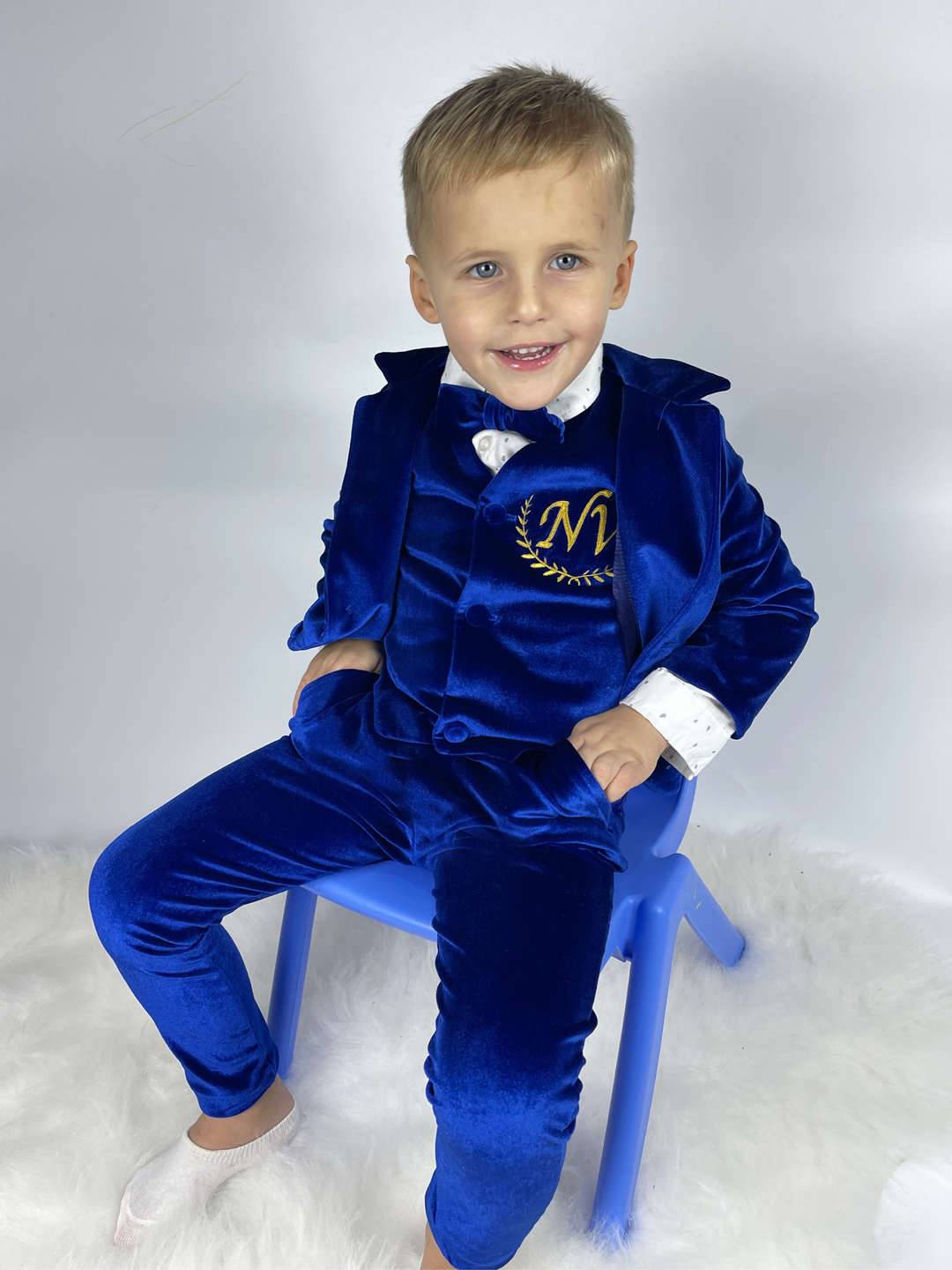 A cheerful 4-year-old boy, with bright blond hair, sits comfortably on a chair. He wears a stylish royal blue tuxedo, complete with a gold-embroidered vest showcasing his initials.