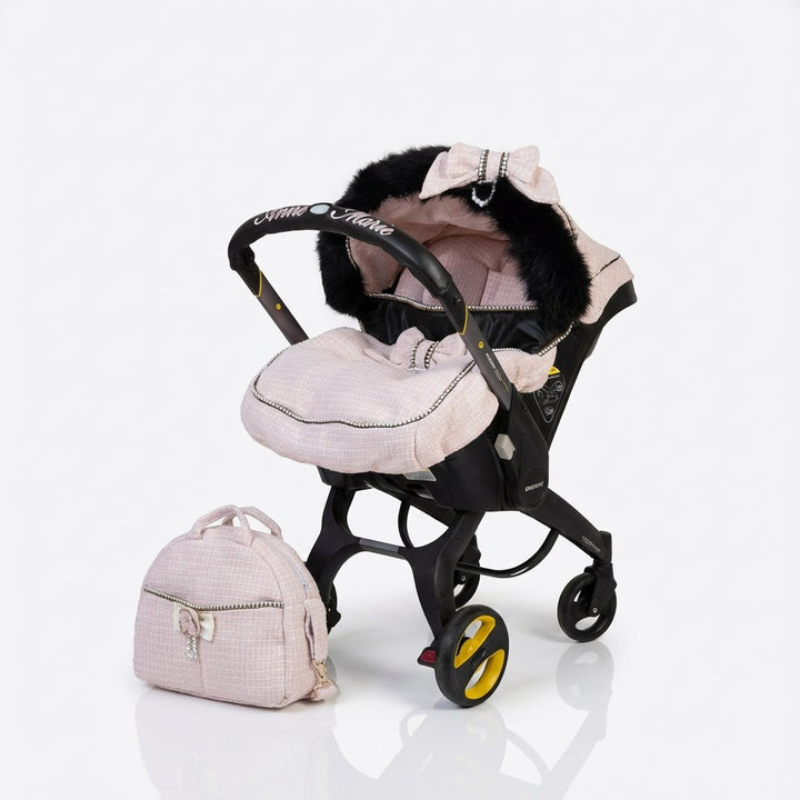 Baby car seat installed on a stroller frame, featuring a light pink tweed fabric set with a Chanel-inspired design, padded footmuff, black real fur lining inside the canopy, embroidery on the carry handle, a light pink bow detail on the hood, and a matching tweed bag placed beside the stroller on a plain light background.