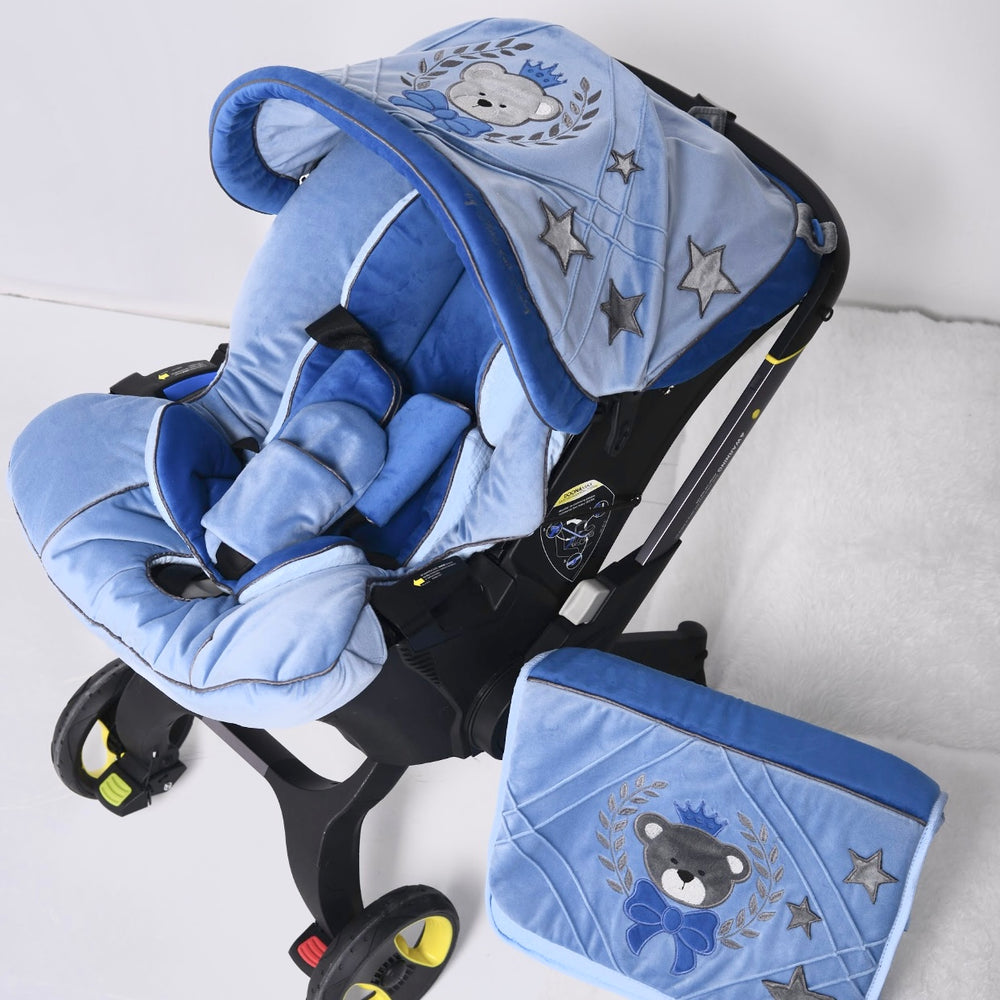 Angled view of a Doona car seat on a light studio background, featuring a light and dark blue velour padded seat insert with contoured head and side supports, a quilted velour canopy with embroidered teddy bear crest, crown and star motifs, rounded canopy edge trim, and a matching quilted velour bag with embroidered teddy bear emblem placed next to the car seat.