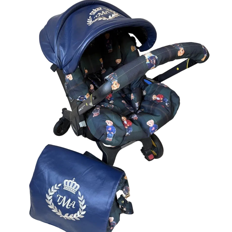 Top-angled view of a Doona car seat installed on the Doona stroller frame. The canopy is made of blue leather with a white embroidered crown and laurel wreath monogram. The seat insert features a dark plaid fabric with Polo-style teddy bear print. A matching blue leather diaper bag with the same embroidered monogram is placed next to the stroller. Shown on a plain light background.