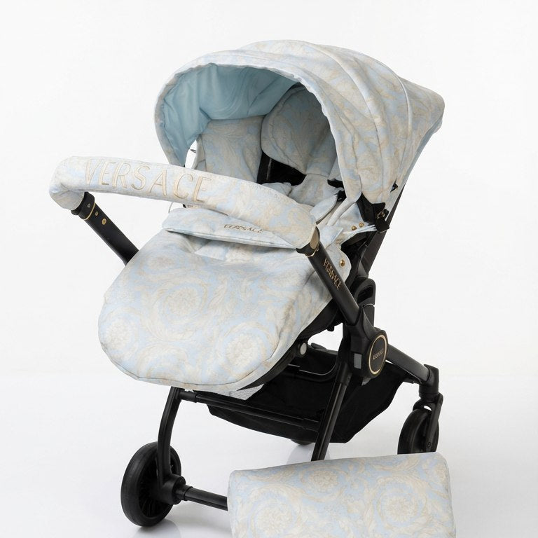 Side view of a baby car seat installed on a black stroller frame, photographed on a white background. The car seat is dressed in a light beige and pale blue fabric set. A rounded canopy with a pale blue interior is extended over the seat. The handlebar cover is light beige and features the word “VERSACE” embroidered in large capital letters across the center. The footmuff matches the canopy and seat cover in the same light beige tone. The stroller wheels and frame are clearly visible.