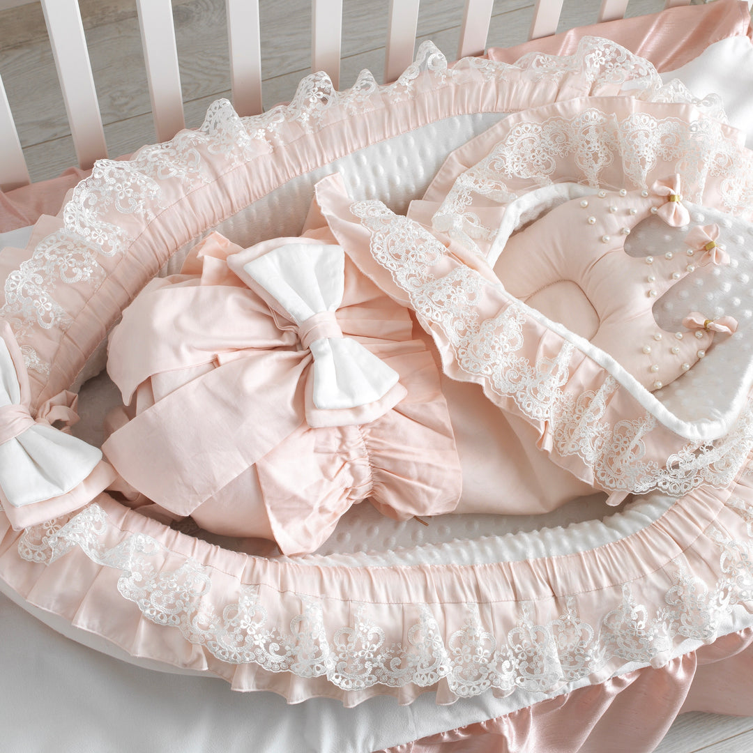 Peach baby nest with white lace details and folded blanket on a white crib