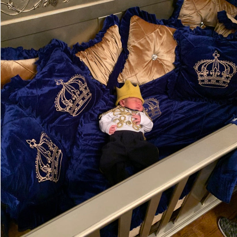 Newborn baby in a crib with royal blue infant bedding and gold crowns, wearing a yellow crown.