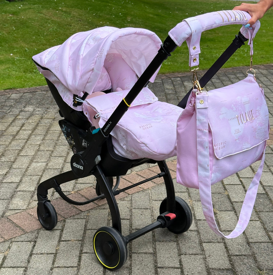 Pink world map car seat cover set on Doona X stroller outdoors on paved walkway with green lawn background, matching diaper bag attached.
