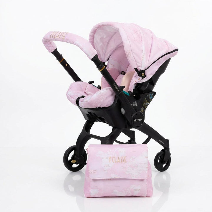 Angled view of a pink world map stroller cover set with gold embroidery, installed on a Doona X car seat, featuring matching seat cover and accessory bag.