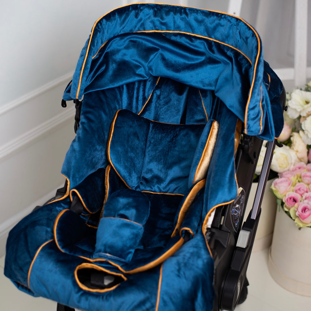 Luxury blue velour baby car seat cover with gold piping, padded inserts and extended canopy, photographed in an elegant interior with floral decor.