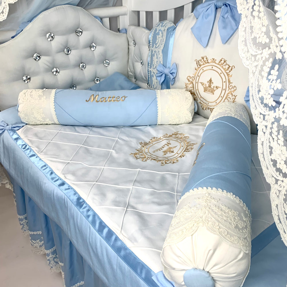 Baby crib bumpers with blue and white bedding, decorative pillows with rhinestones, and roll pillows with name embroidered.