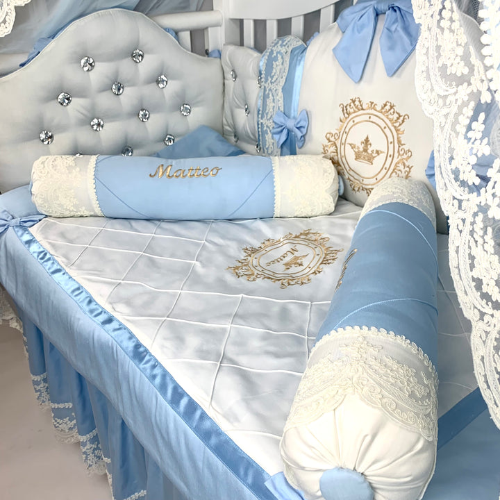 Baby crib bumpers with blue and white bedding, decorative pillows with rhinestones, and roll pillows with name embroidered.
