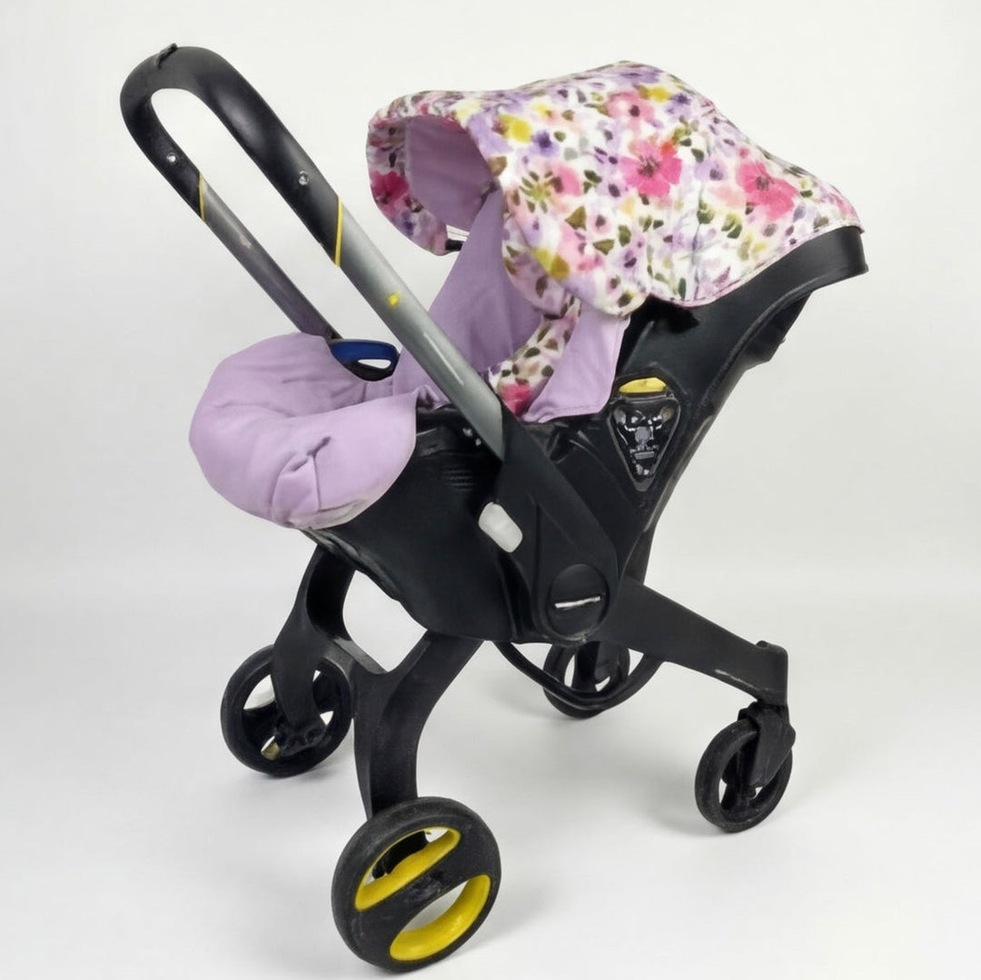Floral baby car seat set installed on a car seat, featuring a soft lavender seat insert and matching shoulder pads, paired with a pastel floral canopy in pink, yellow and green tones with a purple interior lining. Photographed on a clean white background.
