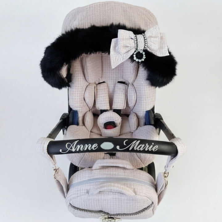 Front view of a baby car seat installed on a stroller frame, upholstered in light pink tweed fabric. The canopy is trimmed with real black fur and decorated with a light pink bow. The carry handle features embroidered text “Anne Marie.” A matching padded footmuff is attached. Displayed on a plain light background.