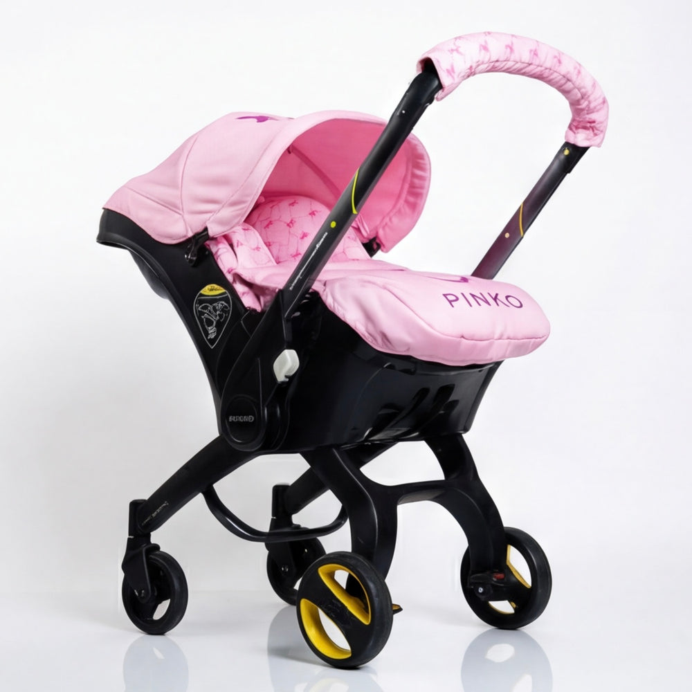 Pink baby car seat set installed on a car seat, featuring a soft pink canopy, padded seat insert, footmuff with Pinko logo, and a matching handlebar cover. Coordinated pink patterned fabric details. Photographed on a clean white background.