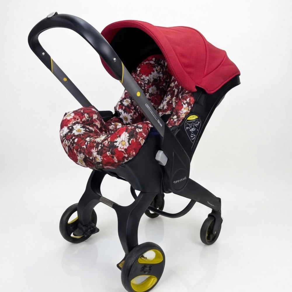 Baby car seat cover installed on a Doona car seat, featuring a red canopy and floral seat padding with a red, white, and dark floral pattern, photographed on a white background.