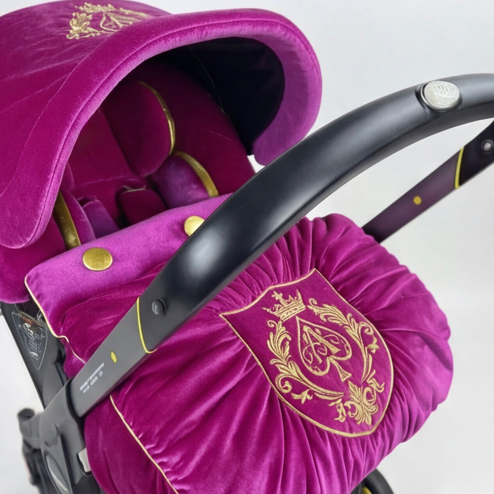 Close-up view of a luxury pink velour baby car seat set with a rich gold interior, gold piping and embroidered crest, installed on a Doona car seat and photographed on a white background.