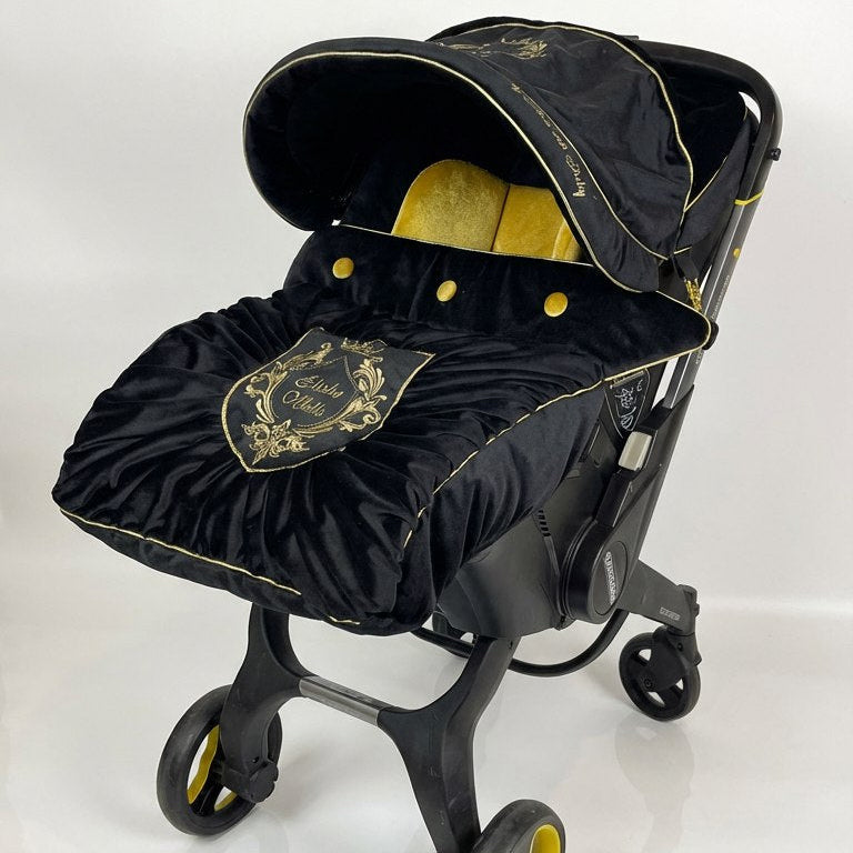 Black velvet Doona+ car seat cover set with gold piping, gold embroidered crest and gathered velvet footmuff with gold buttons, shown on stroller frame.
