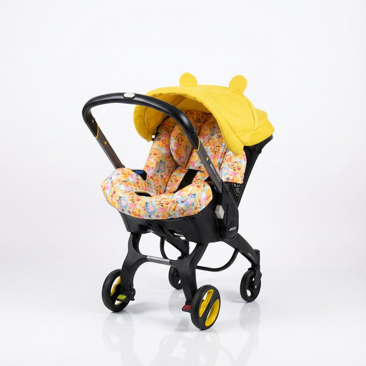 Doona car seat installed on the Doona stroller frame, shown on a light background. The set features a bright yellow canopy with rounded ear details and a seat insert with a Winnie the Pooh–inspired multicolor print. Black stroller frame with integrated wheels visible.