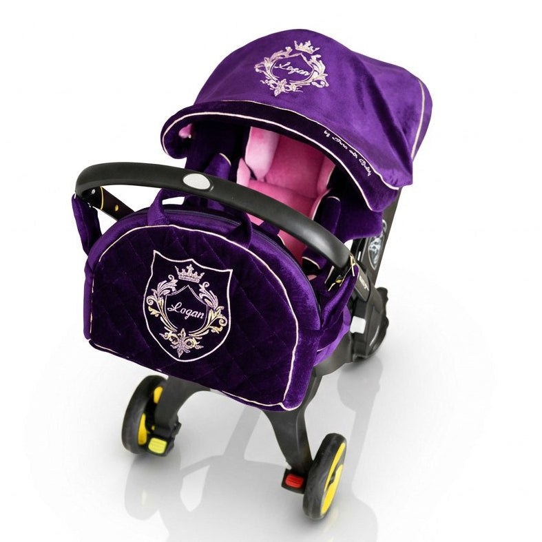 Purple velvet baby car seat cover set installed on a car seat on a white background, featuring a padded canopy with white crest embroidery, a matching footmuff and seat cover with personalized name embroidery, and elegant contrast piping.