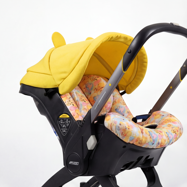 Side view of a Doona car seat installed on the Doona stroller frame, shown on a light background. The setup features a yellow canopy with rounded ear details and a seat insert with a Winnie the Pooh–inspired multicolor print. Black frame and integrated wheels visible.