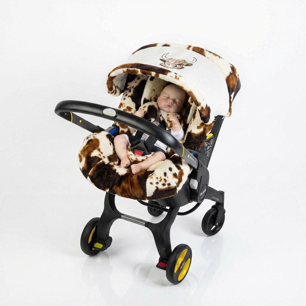 Plush baby car seat cover with a brown and cream cow-print design, featuring soft padded inserts and an embroidered bull head on the canopy. Shown with a sleeping baby inside, photographed on a clean light studio background.