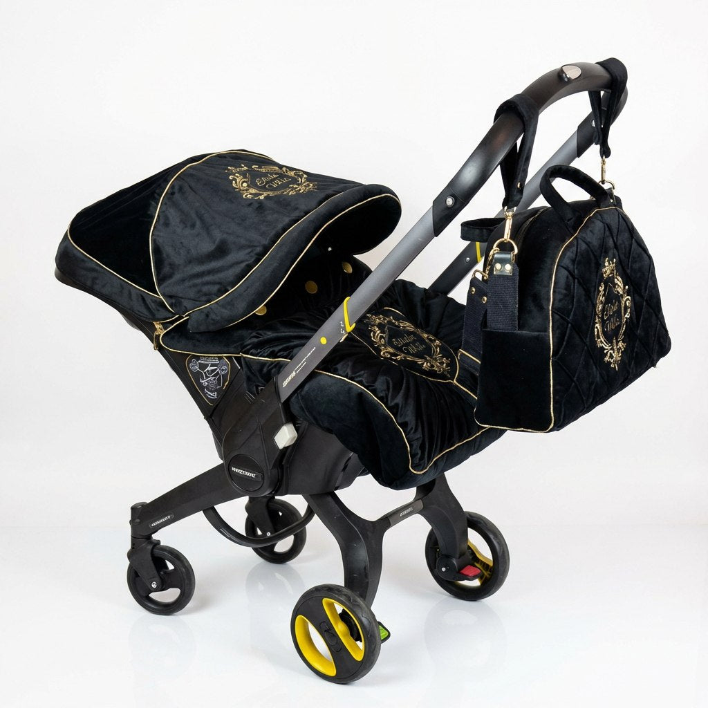 Black infant car seat cover set with gold piping and embroidered crest details, including a matching canopy, padded seat cover, and coordinating black bag attached to the handle, shown on a wheeled infant car seat frame against a white background.