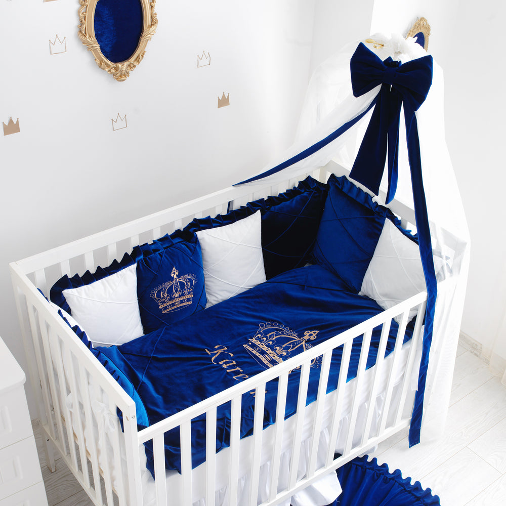 White crib with blue baby bedding set featuring crowns and 'Kings' text, in a room with decorative elements.