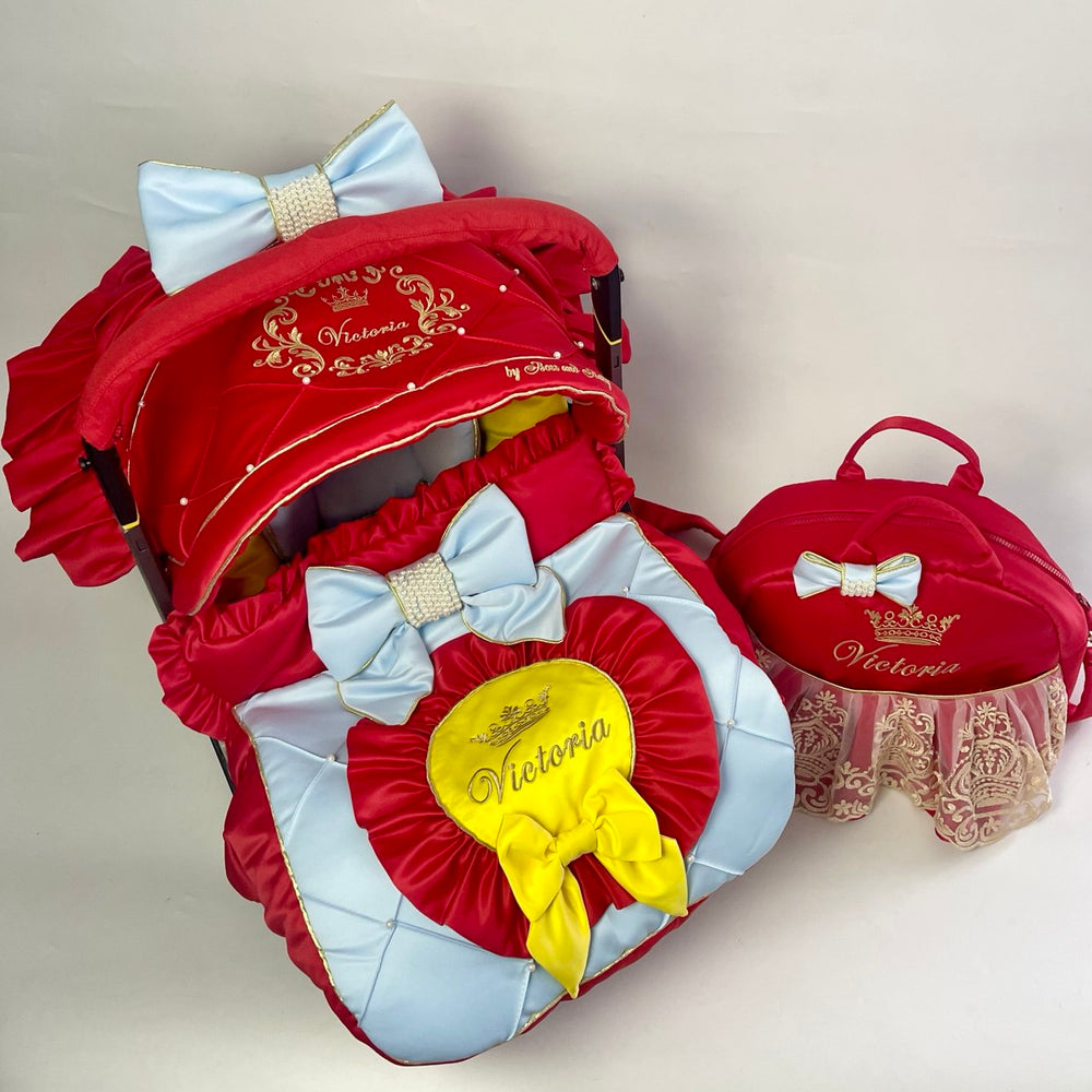 Red and light‑blue car seat cover set with satin quilting, gold “Victoria” embroidery, blue bows with pearl center  and yellow bow; white studio background.
