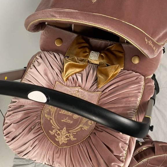 Blush pink velvet Doona car seat cover set with gold trimming, pleated footmuff, embroidered crest and decorative gold velvet bow.