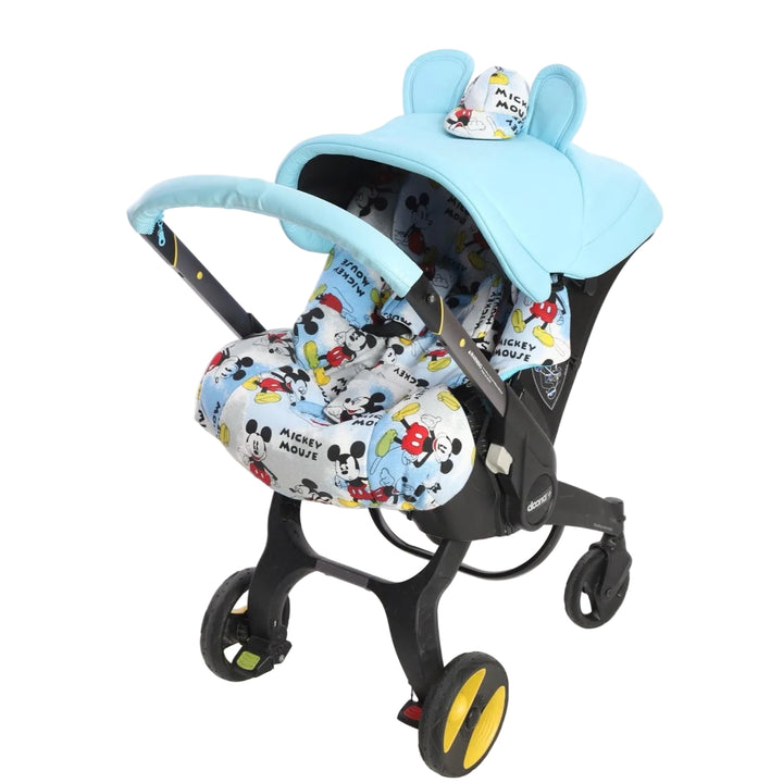 Light blue baby car seat cover set installed on a car seat with stroller wheels, photographed on a white background. The canopy is light blue with rounded shape and decorative bunny ears on top, featuring a small printed patch with Mickey Mouse characters. The seat cover and foot area have an all-over Mickey Mouse print in black, red, yellow, and light blue on a white background. The handlebar is covered with a matching light blue fabric cover. The car seat is shown from a front three-quarter angle.