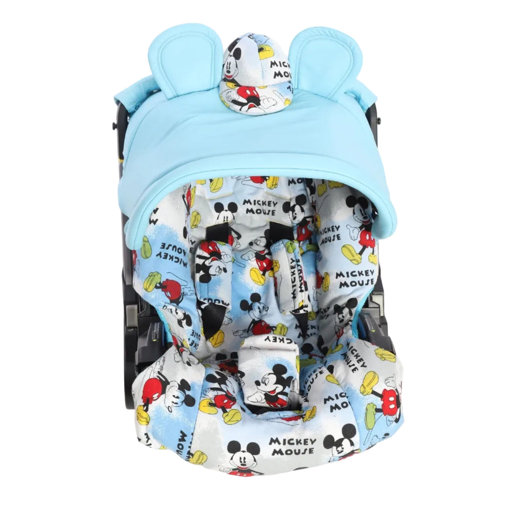 Front view of a baby car seat with a light blue canopy installed, photographed on a white background. The canopy has a rounded shape with two decorative bunny ears and a small central patch made of Mickey Mouse printed fabric. Inside the car seat, the seat liner, shoulder pads, and crotch pad feature an all-over Mickey Mouse print in black, red, yellow, and light blue on a white background. The car seat frame is partially visible on both sides.