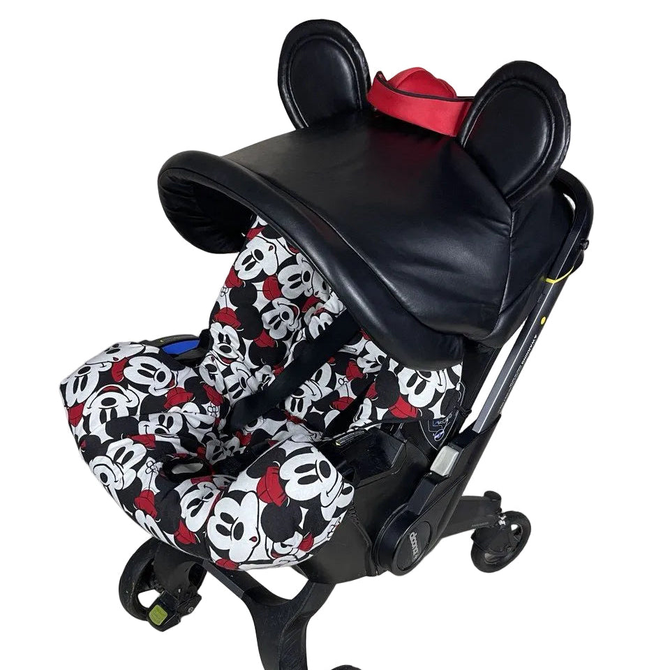 Black baby car seat mounted on a stroller frame, featuring a black leather-look canopy with rounded ear details and a red hat-style accent on top, paired with a black, white, and red Mickey Mouse print seat cover, photographed on a plain light background.