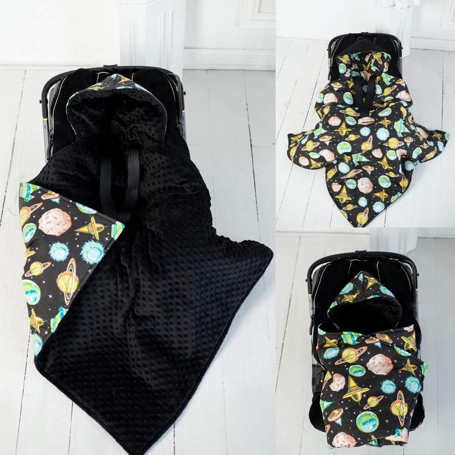 Hooded baby blanket transforming into an envelope-style wrap, shown in multiple views. The left image shows the blanket partially opened with a black textured interior and a space-themed outer fabric, while the right images show the blanket fully wrapped around a baby car seat. The outer fabric features a colorful space print with planets, stars, and rockets, photographed on a light interior background.
