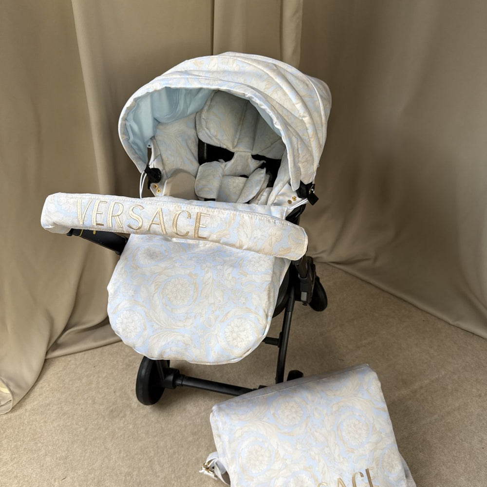 Baby car seat installed on stroller, beige Versace-inspired set with jacquard pattern. Matching hood and footmuff in light beige tones, handlebar cover with gold “VERSACE” embroidery. Placed on a neutral beige fabric background, matching bag visible in front.