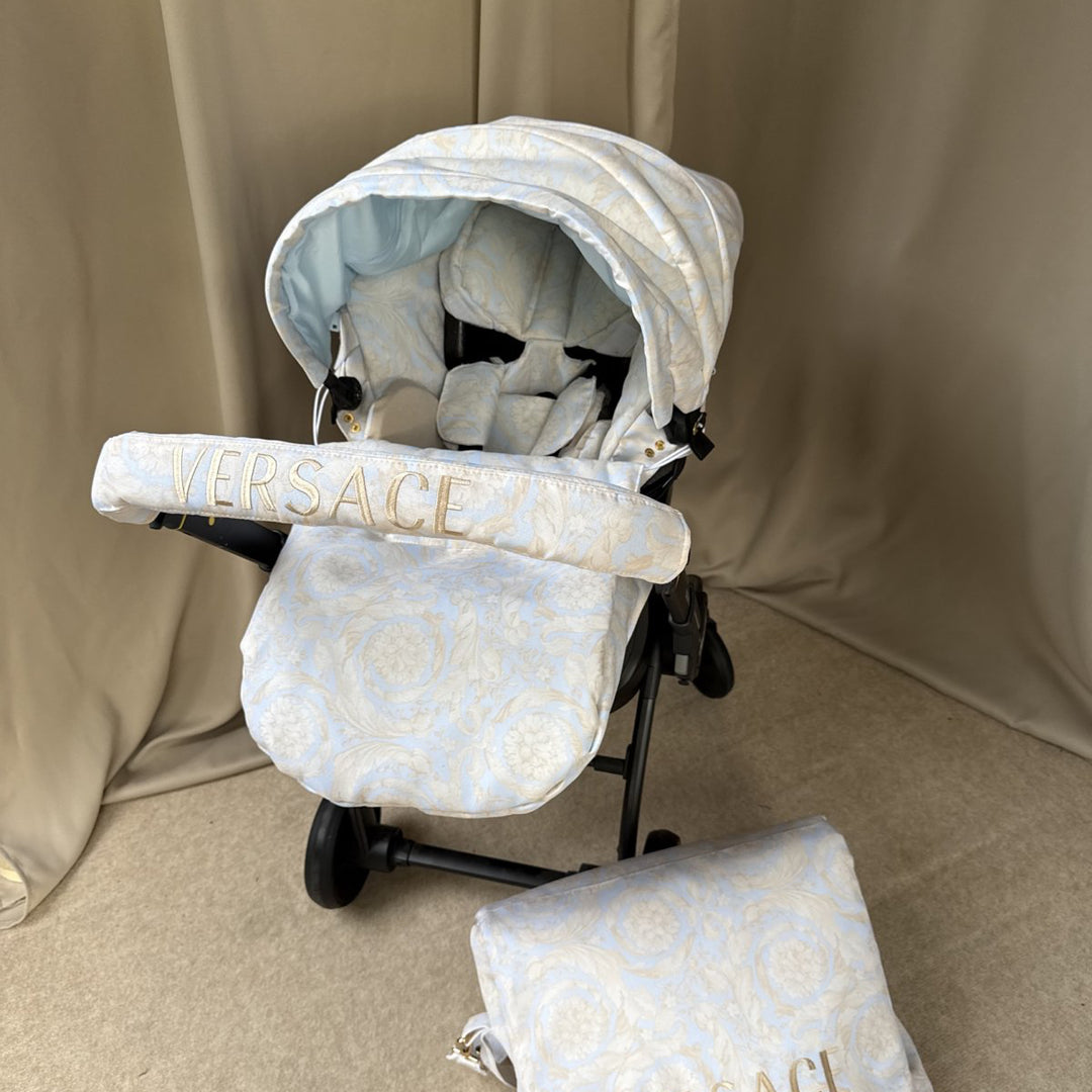Baby car seat installed on stroller, beige Versace-inspired set with jacquard pattern. Matching hood and footmuff in light beige tones, handlebar cover with gold “VERSACE” embroidery. Placed on a neutral beige fabric background, matching bag visible in front.